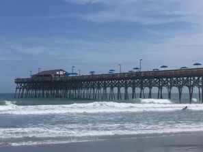 Un muelle que se adentra en el agua, con un edificio al final y sombrillas sobre él.