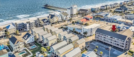 A coastal residential area with a pier extending into the ocean, featuring a mix of single and multi-story houses, a parking lot, and a swimming pool area.