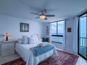 A bedroom with a bed, nightstand, lamp, and a view of the sea through a large window. - Artsy Waterfront Retreat - Punta Rassa 4-1003 (Fort Myers)