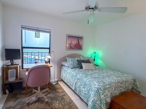 Ein Schlafzimmer mit Bett, Schreibtisch, Stuhl, Fenster mit Blick, Deckenventilator und einer Wandleuchte.