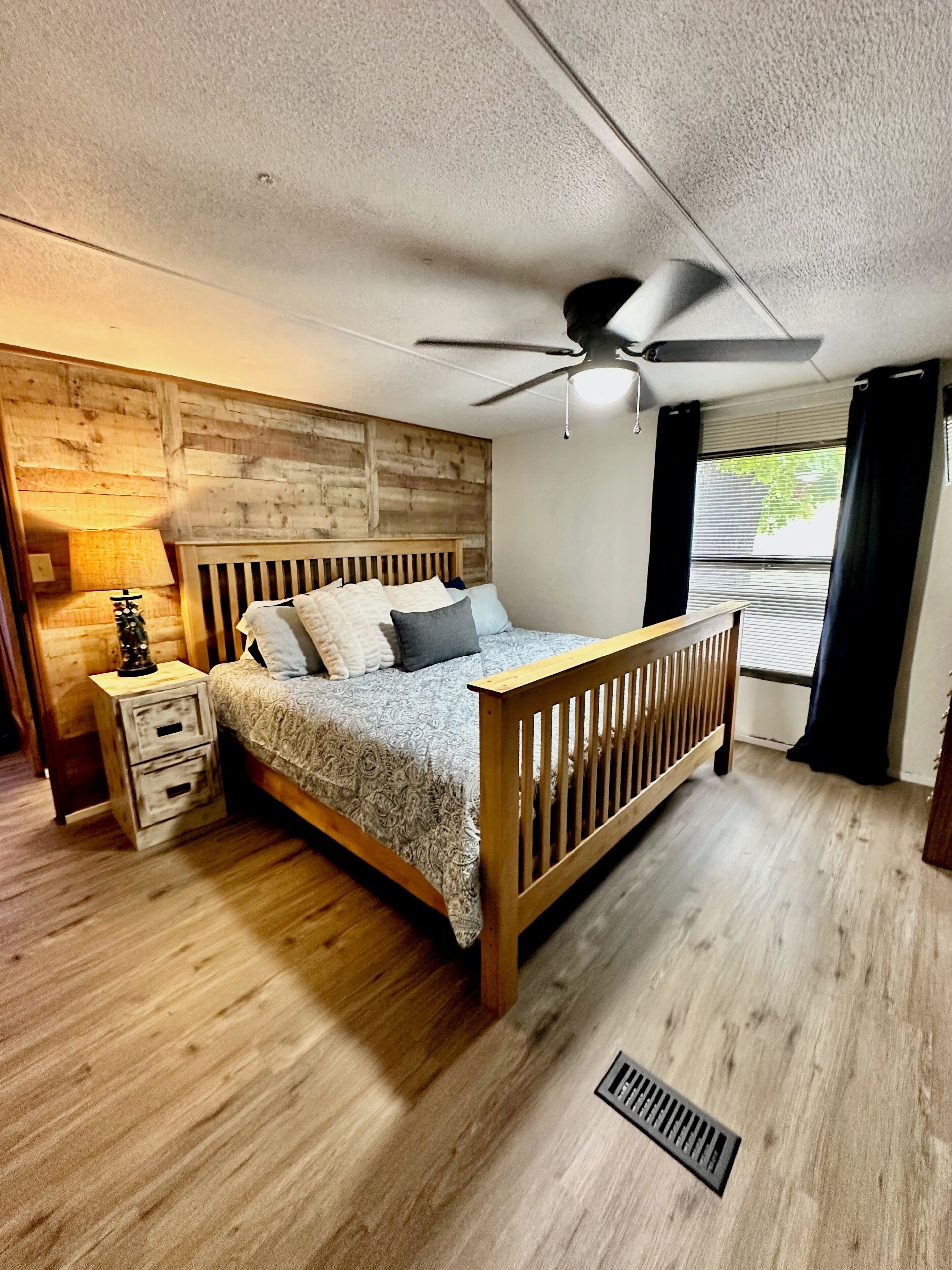 A bedroom with a wooden headboard, a bed with patterned bedding, a nightstand with a lamp, and a ceiling fan.