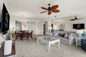 A spacious living room with a large sofa, a glass coffee table, and a dining area with a chandelier. - New! Gulf Front! Pool, Hot Tub, Steps to the Beach, Beach Manor 210 (Miramar Beach)