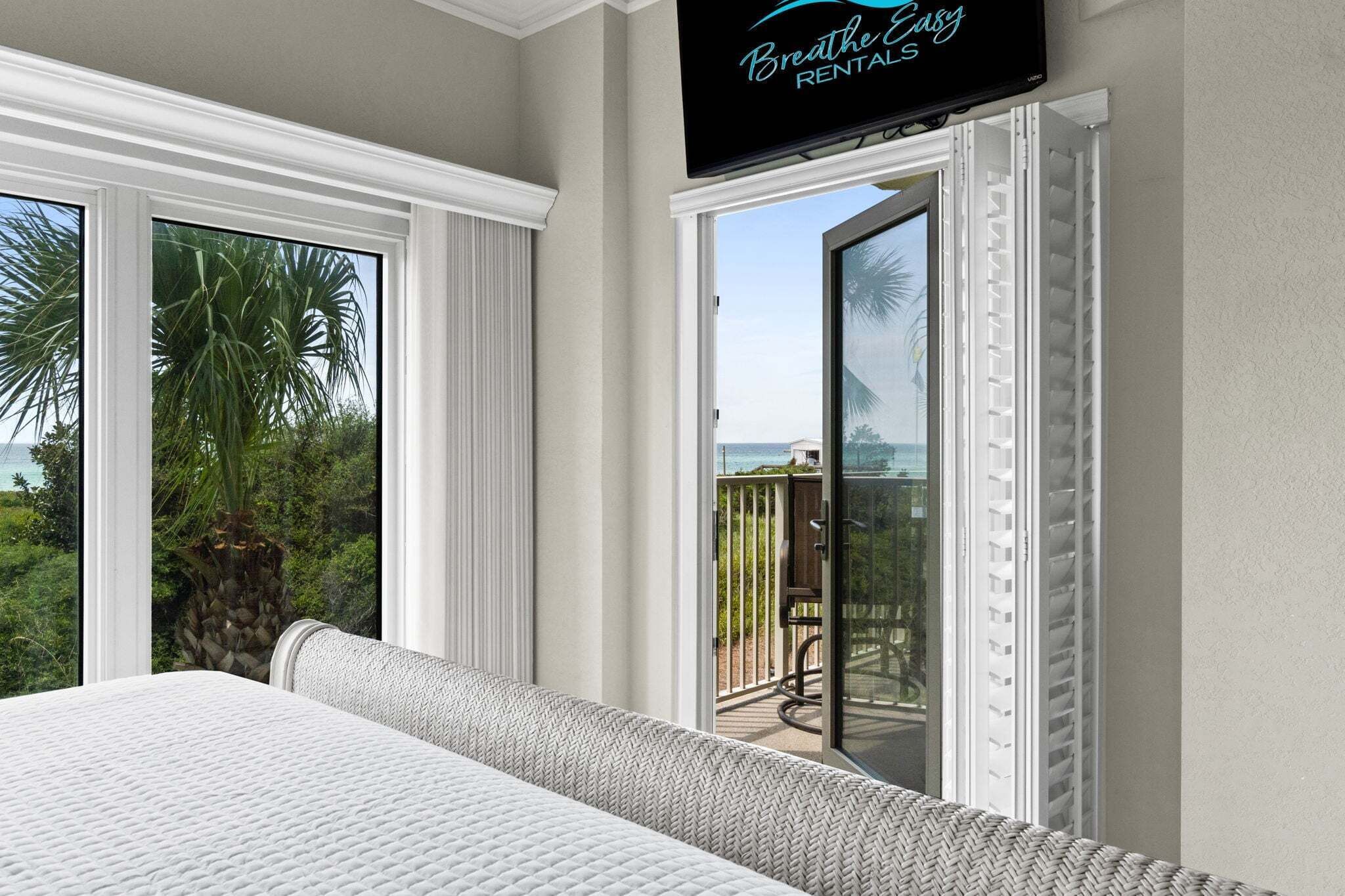 A bedroom with a large bed, a television mounted on the wall, and a sliding glass door leading to a balcony with a view of the ocean.