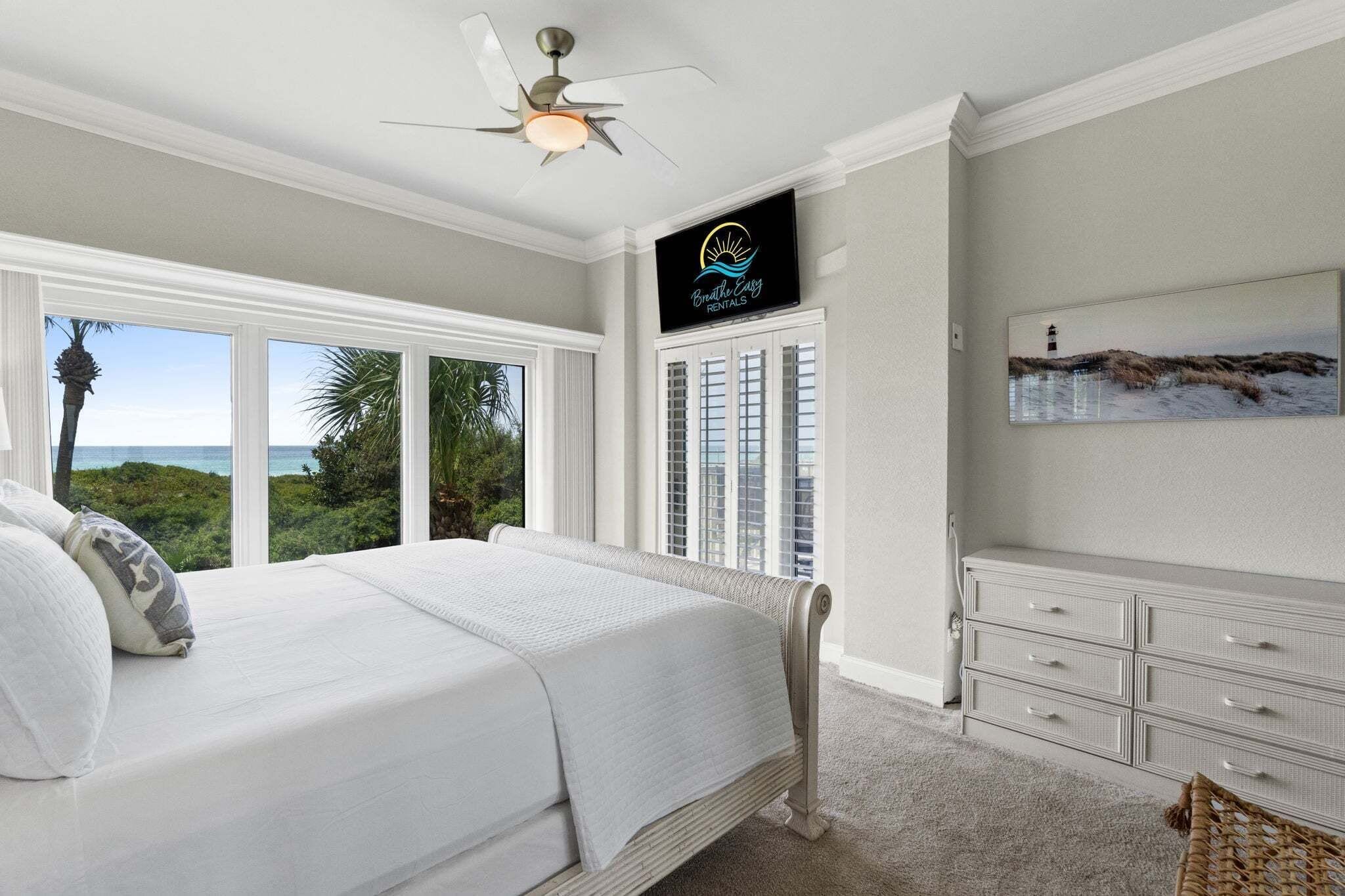 A bedroom with a large bed, a ceiling fan, a view of the ocean, a sign for 'Breathe Easy Rentals', and a dresser.