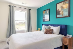 Ein Schlafzimmer mit einem großen Bett, einem Fenster mit Vorhängen, einem Nachttisch und zwei Wandbildern.