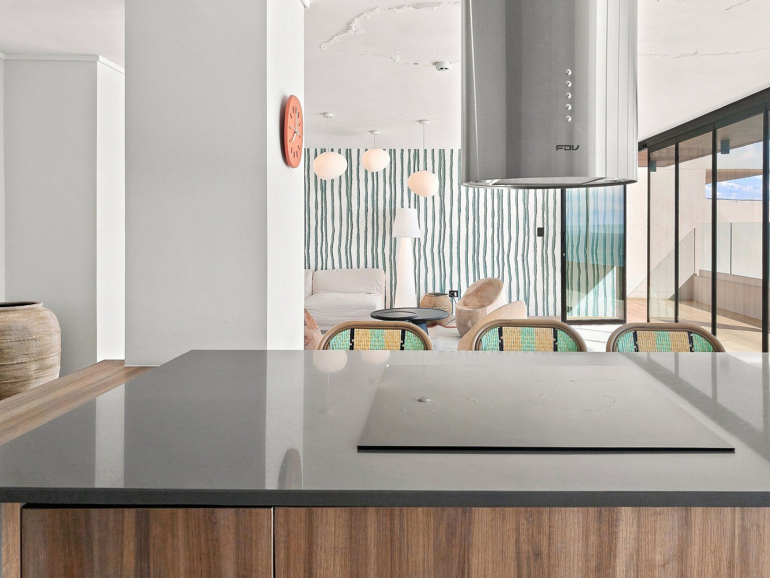 A modern kitchen with a large island, wooden cabinets, and a view of a dining area with striped wallpaper and a sofa.