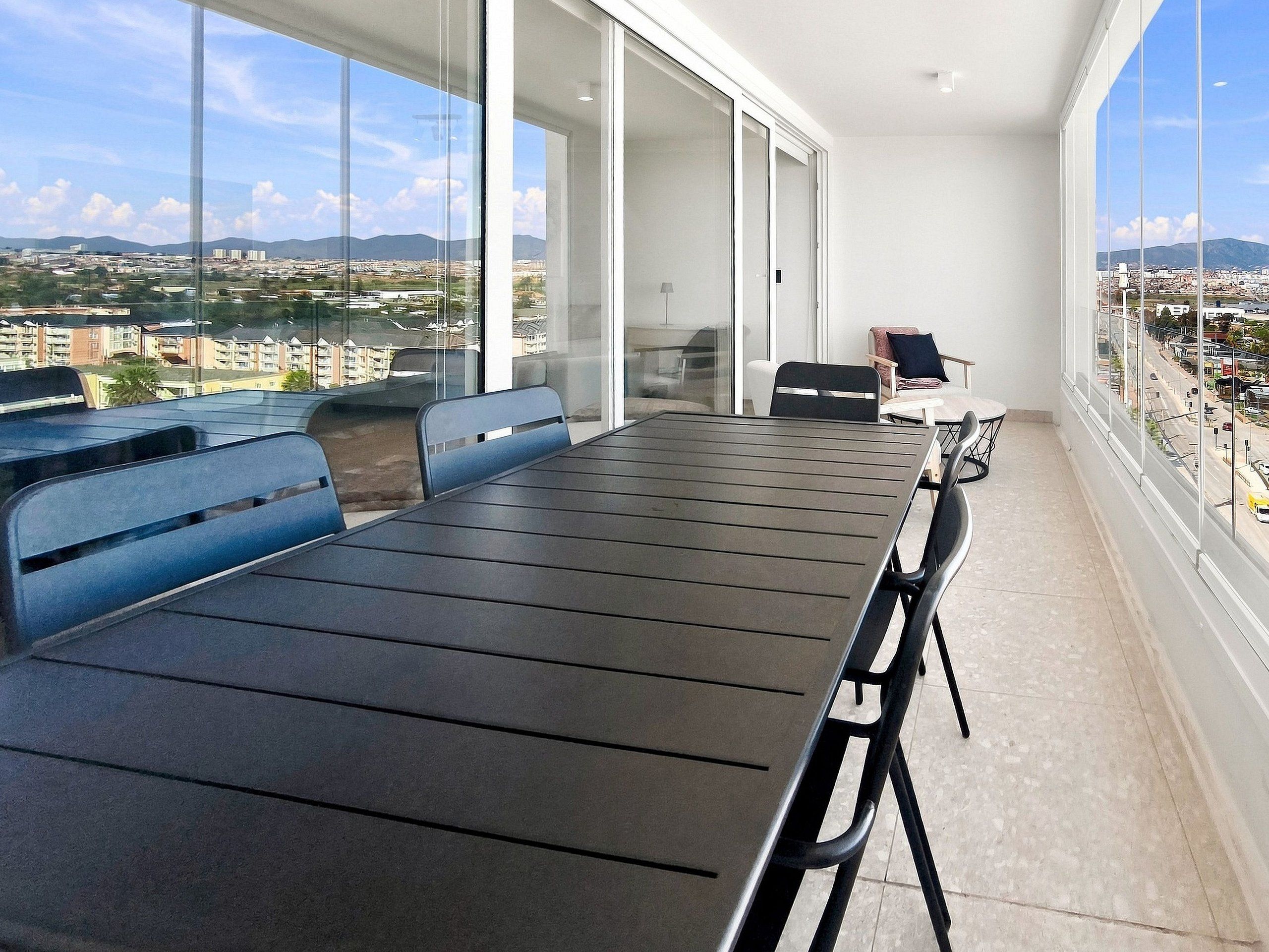 A modern balcony with a dining table and chairs, large windows offering a city view, and a sofa.