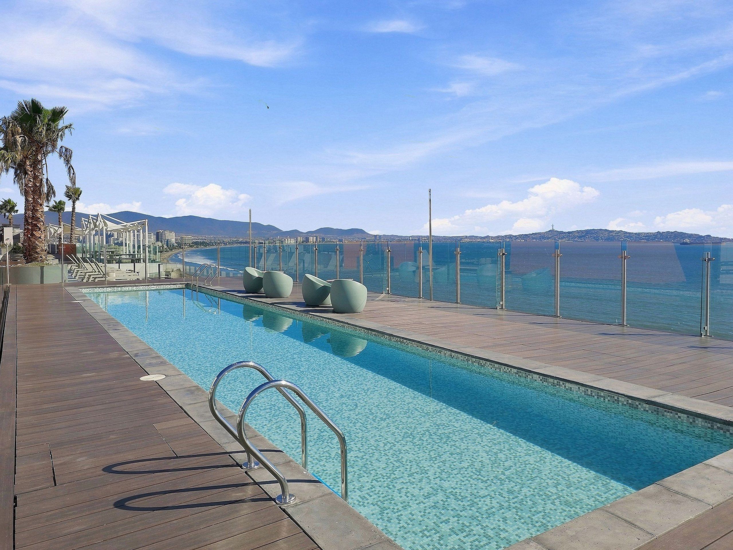 A swimming pool with a view of the sea and mountains, surrounded by palm trees and modern seating.