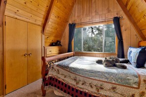 A bedroom with a wooden ceiling, a large window with curtains, a bed with a patterned blanket, and a wooden dresser. - Mountain Getaway Near Snow Summit | 4BR • 3.5BA • Game Room (Big Bear Lake)