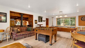 A cozy room with a foosball table, a billiards table, and a large window offering a view of the outdoors. - Mountain Getaway Near Snow Summit | 4BR • 3.5BA • Game Room (Big Bear Lake)