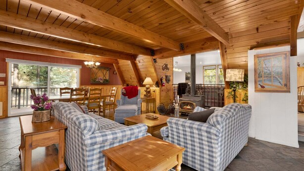 A cozy cabin interior with a wooden ceiling, a fireplace, checkered sofas, and a dining area. - Mountain Getaway Near Snow Summit | 4BR • 3.5BA • Game Room (Big Bear Lake)