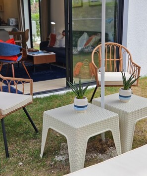 Ein Sitzbereich im Freien mit Korbstühlen, Tischen, Pflanzgefäßen und Blick ins Innere durch eine Glastür.