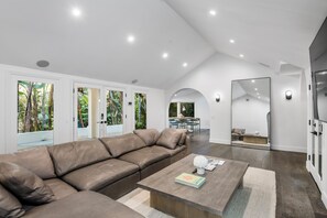 Een moderne woonkamer met een grote bruine leren bank, een houten salontafel, en een eethoek zichtbaar door een boogvormige doorgang.
