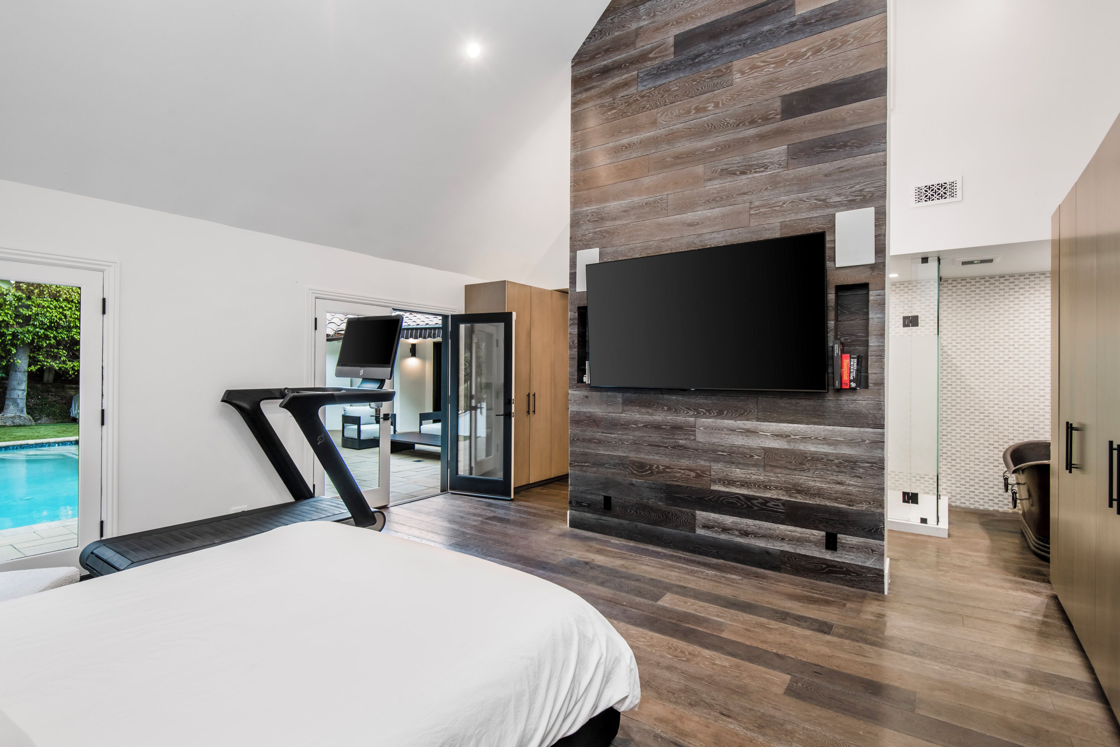Un dormitorio moderno con una cama grande, un televisor montado en una pared de madera y un ventanal corredizo que da a un balcón.