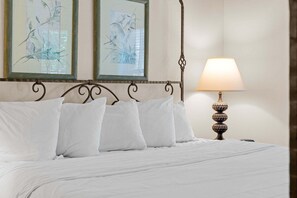 Ein ordentlich bezogenes Bett mit weißen Leinen und Kissen, eine Wandlampe sowie zwei gerahmte Kunstwerke darüber.