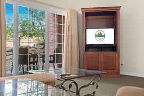 A room with a glass table, a wooden cabinet with a television, and a view of a brick wall and greenery outside. - Relax at French Quarter | 2BR Condo with Balcony (Branson)