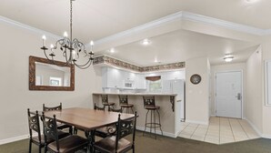 Uma sala de jantar com mesa e cadeiras de madeira, um candelabro e uma cozinha ao fundo.