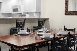 Uma sala de jantar com uma mesa de madeira posta para quatro pessoas, cercada por cadeiras pretas com estampas florais. Pratos e copos brancos estão dispostos na mesa.
