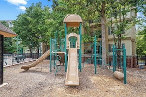 Ein Spielplatz mit Rutsche und Klettergerüsten, umgeben von Bäumen und einem Zaun.