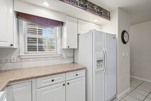 A kitchen with white cabinets, a refrigerator, a window with blinds, and a clock on the wall. - Prime Branson Getaway | Near the Strip & Shows (Branson)