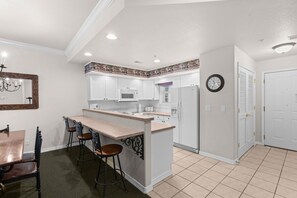 A kitchen with white cabinets, a microwave, a refrigerator, a clock, and a dining area with a table and chairs. - Stay in the Heart of Branson | French Quarter (Branson)
