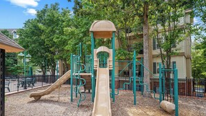 Un parque infantil con tobogán y estructuras para escalar, rodeado de árboles y una valla.