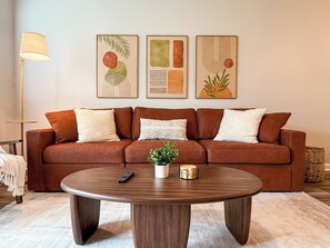 Ein Wohnzimmer mit einem braunen Sofa, einem runden Holztisch und drei gerahmten Kunstwerken an der Wand.