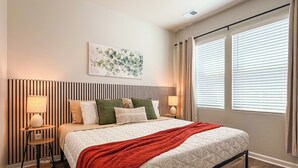 A bedroom with a bed, bedside lamps, a headboard with vertical lines, a window with blinds, and a wall-mounted artwork. - Modern 3BR/3BA Family Home - Beach • Pool • Shops (Myrtle Beach)