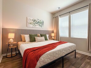 A bedroom with a bed, bedside lamps, a headboard with vertical lines, a window with blinds, and a wall-mounted artwork. - Modern 3BR/3BA Family Home - Beach • Pool • Shops (Myrtle Beach)