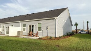A single-story residential building with a grey exterior, a covered patio with wooden chairs, and a well-maintained lawn. - Modern 3BR/3BA Family Home - Beach • Pool • Shops (Myrtle Beach)