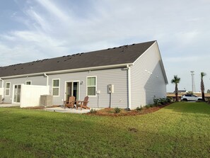 A single-story residential building with a grey exterior, a covered patio with wooden chairs, and a well-maintained lawn. - Modern 3BR/3BA Family Home - Beach • Pool • Shops (Myrtle Beach)
