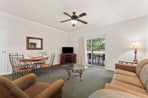 Ein Wohnzimmer mit Deckenventilator, einem Esstisch mit Stühlen, einer Couch, einem Fernseher auf einer Anrichte und Blick durch eine Glastür.