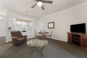 A living room with a glass-top table, a brown sofa, a ceiling fan, a television, and a kitchen in the background. - Pet Friendly Queen Condo | French Quarter Resort (Branson)