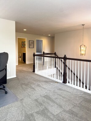 A carpeted hallway with a white railing, a black office chair, and a hanging light fixture. - Lovely 4-bedroom house in Bright and Charming Brighton Next to Pool and Gym (Brighton)