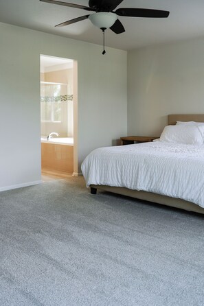 Une chambre à coucher avec un lit, un ventilateur de plafond et une salle de bain visible à travers une porte ouverte.