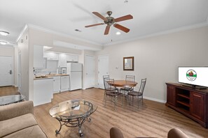 A spacious living room with a glass coffee table, a dining area with a wooden table and chairs, a kitchen with white cabinets, and a ceiling fan. - Queen Condo Near Shopping & Entertainment (Branson)