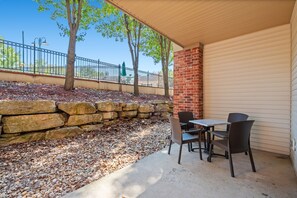 An outdoor patio with a stone wall, a table with chairs, and a view of a fenced area with trees. - Queen Condo Near Shopping & Entertainment (Branson)