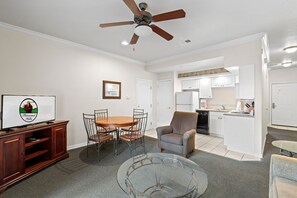 A living room with a wooden dining table, chairs, a glass coffee table, a sofa, a television, and a ceiling fan. - Stay Close to the Strip | Pet Friendly Queen Condo (Branson)