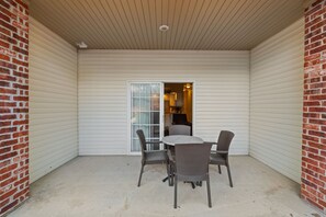 Un'area patio coperta con tavolo e sedie, una porta scorrevole in vetro che dà su una cucina e un muro in mattoni su un lato.