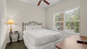Ein ordentlich bezogenes Bett in einem Zimmer mit Deckenventilator, einem kleinen Tisch mit Lampe und einem Fenster mit Jalousien.