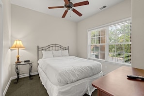 A neatly made bed in a room with a ceiling fan, a small table with a lamp, and a window with blinds. - Stay Close to the Strip | Pet Friendly Queen Condo (Branson)