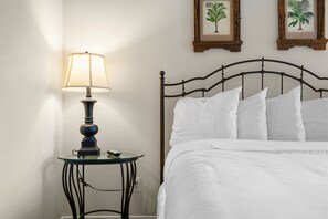 A bed with white bedding, a metal headboard, and a bedside table with a lamp. There are two framed botanical prints on the wall. - Queen Condo Near Highway 76 | Pool & Hot Tub (Branson)