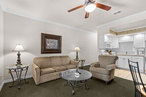 Ein Wohnzimmer mit einer Couch, einem Sessel, einem Couchtisch und zwei Lampen. Im Hintergrund ist ein Deckenventilator und eine Küchenzeile zu sehen.