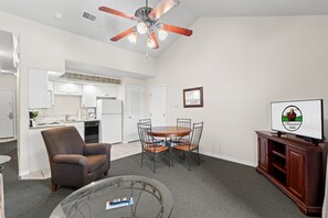 A living room with a brown armchair, a glass coffee table, a dining table with chairs, a ceiling fan, a kitchen area, and a television on a wooden stand. - Resort Queen Condo | Balcony • Pool • Hot Tub (Branson)