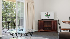 A room with a glass table, a television on a wooden cabinet, and a view of greenery outside. - Relax at French Quarter Resort | Queen Condo (Branson)