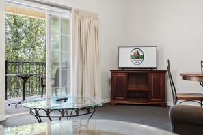 A room with a glass table, a television on a wooden cabinet, and a view of greenery outside. - Relax at French Quarter Resort | Queen Condo (Branson)