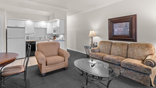 Una sala de estar moderna con sofá, sillón y mesa de centro, una cocina con armarios blancos y un ventilador de techo.