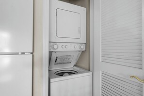 A compact, white laundry room with a built-in washer and dryer, a control panel, and a door with horizontal blinds. - Relaxing King Condo | Close to Shows & Attractions (Branson)