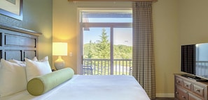 Habitación de hotel con cama, televisión, lámpara y balcón con vistas a árboles.