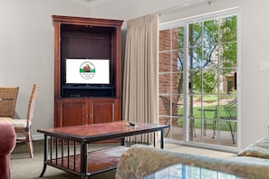 Ein Zimmer mit Fernseher, einem Holzkasten, einem Tisch und Blick auf eine Terrasse mit Gartenmöbeln.